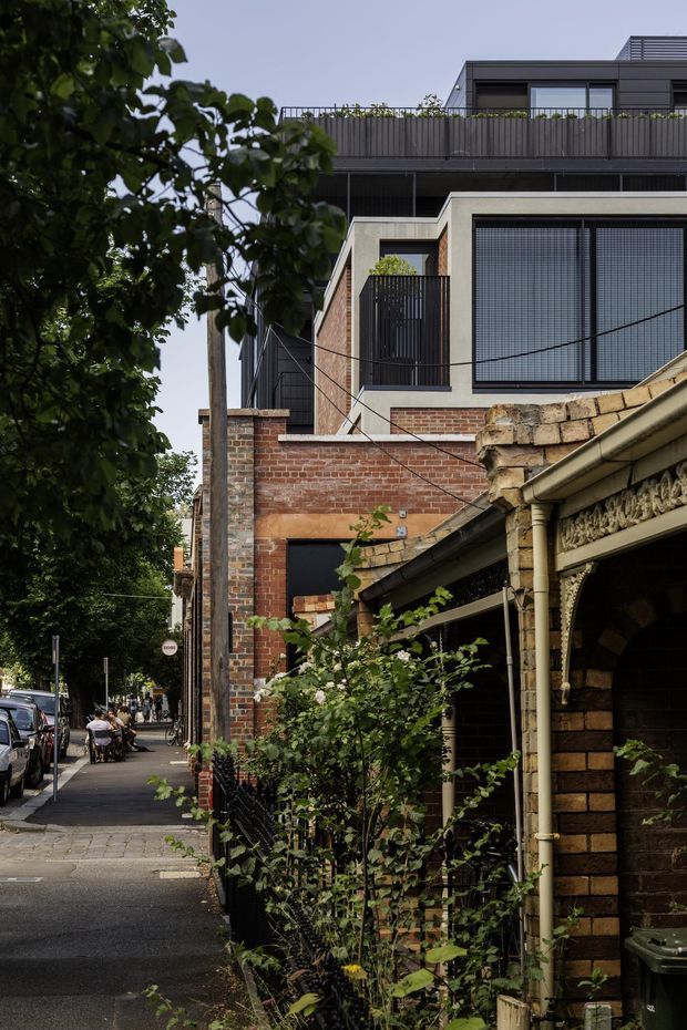 Street-level exterior view of a dense urban residential setting, showing a contemporary multi-storey extension rising behind older brick terrace-style buildings. The newer structure has dark cladding, flat rooflines, large gridded windows, balconies and planted roof terraces, while the foreground retains ornate historic brickwork and a traditional covered entry. Trees, footpaths and parked cars frame the streetscape.