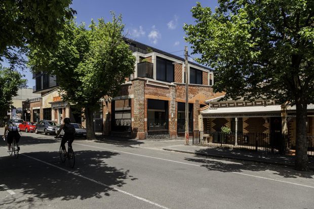 Street-level exterior view of a multi-storey urban building with exposed red-brick walls, dark-framed glazing, projecting upper-level forms, and a sheltered ground-floor frontage. Mature street trees, parked vehicles, cyclists, and neighbouring façades frame the scene.