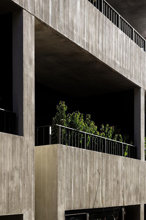 Close-up exterior view of a strongly geometric concrete building facade, showing tall textured board-formed concrete walls, deep recessed balcony or terrace openings, dark shadowed voids, and slim black metal railings. Sparse greenery is visible within the recessed terrace. The composition emphasizes heavy massing, vertical texture, and high contrast between concrete and shadow.