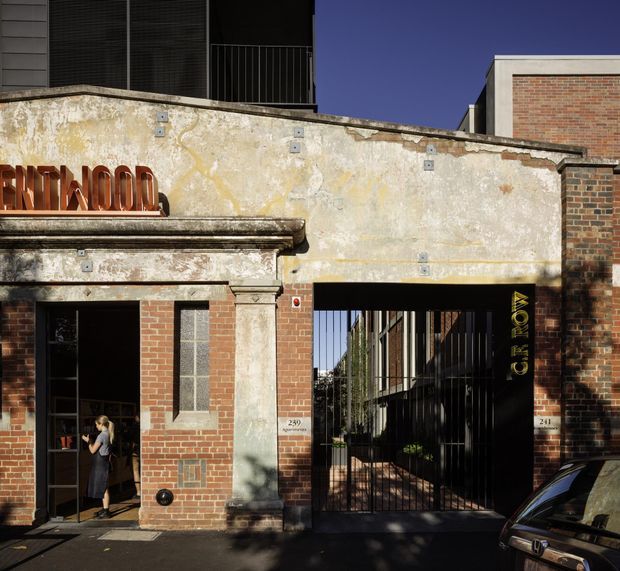 Street-level view of an urban heritage-style brick facade with weathered rendered masonry, a classical stone-trimmed entrance, and a gated passage leading into a contemporary courtyard or development. Dark metal window and gate elements contrast with the aged cream facade and red brickwork. Partial signage and building numbers are visible, with a parked car at the edge of the frame.