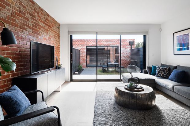 Contemporary living room with an exposed red-brick feature wall, light flooring, a large grey sectional sofa, textured rug, and a rustic round timber coffee table. Full-height sliding glass doors open to a private brick courtyard, while black-framed glazing, modern furniture, and minimalist built-in cabinetry create a refined urban interior.