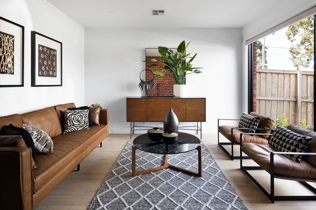 A bright, contemporary living room with a tan leather sofa and matching leather lounge chairs arranged around a round black coffee table. A geometric grey rug, patterned cushions, framed wall art, timber sideboard, indoor plants, and large sliding glass doors create a warm, stylish interior with a view to a fenced outdoor area.