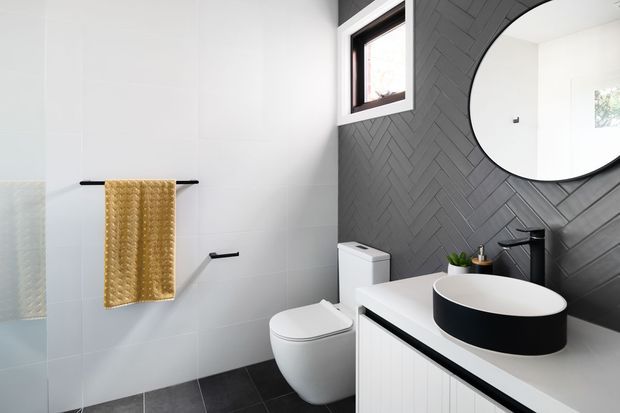 Contemporary bathroom featuring a white tiled wall, dark grey herringbone feature wall, round black-framed mirror, wall-mounted black tapware, black vessel basin on a white vanity, toilet, dark floor tiles, and a mustard textured towel.
