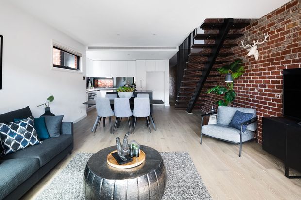 Open-plan contemporary living, dining and kitchen area with a dark sofa, dining table and chairs, compact white kitchen, exposed red-brick feature wall, and a striking black metal open-riser staircase leading to an upper level. Light timber-look flooring and a neutral palette are balanced by dark furniture and blue accents.