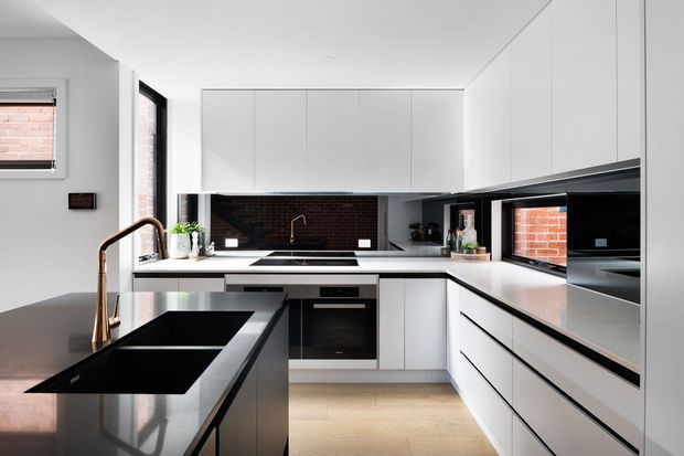 A contemporary white kitchen with an L-shaped layout and central island, featuring flat-panel white cabinetry, dark benchtops, a black brick splashback, integrated appliances, and a bronze-toned gooseneck tap. Large windows and a skylight-like bright ceiling bring natural light into the space.
