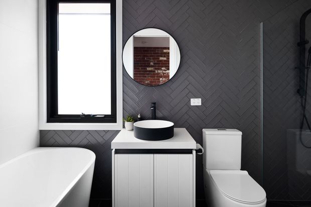 A compact contemporary bathroom with a freestanding white bathtub, wall-mounted black-framed window, dark charcoal herringbone wall tiles, round mirror, white vanity with black vessel basin, and white toilet. The high-contrast monochrome palette creates a refined, dramatic look.