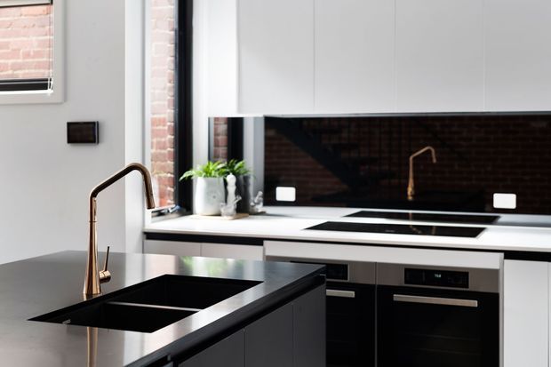 Contemporary kitchen with a dark island featuring an undermount double-bowl sink and brass-toned gooseneck tap. White cabinetry and benchtops contrast with a glossy black splashback, integrated cooktop, and built-in black oven. Exposed brick is visible through the window and reflected in the splashback, while a small potted plant adds greenery.