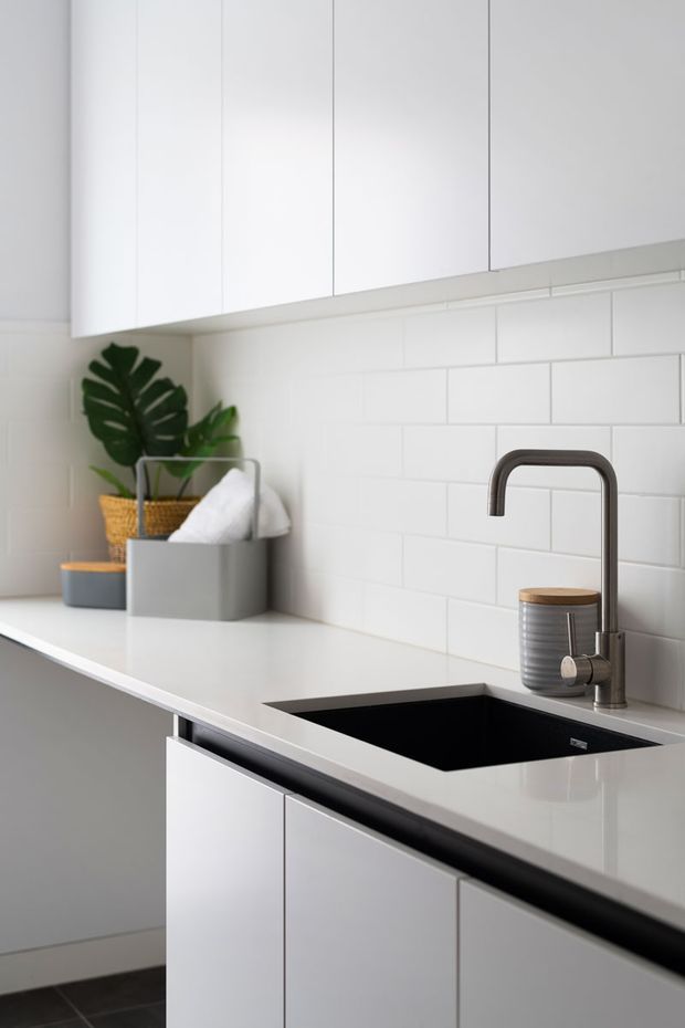 A close-up view of a clean, minimalist kitchen featuring white flat-panel cabinetry, a white engineered-stone countertop, white subway-tile splashback, and a deep black undermount sink with a dark metal gooseneck tap. A small plant, tissue box, and canister provide limited decorative detail.