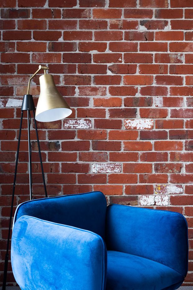 A close-up interior vignette featuring a vivid blue upholstered armchair in front of a red exposed-brick wall. A slender black-and-brass floor lamp with a conical shade stands behind the chair, creating a bold, colourful, slightly industrial living-space composition.