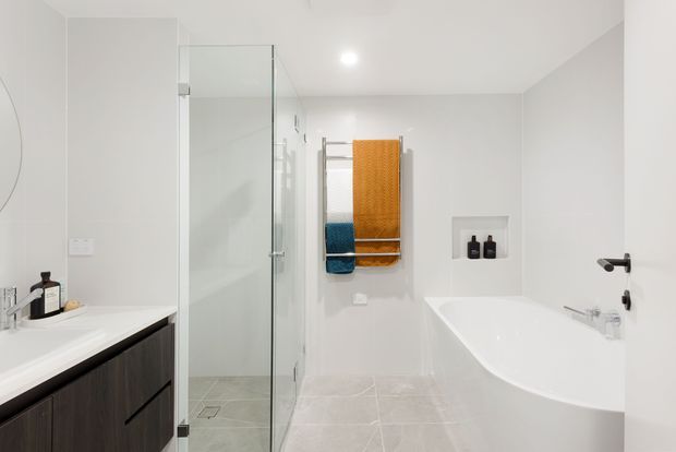 Bright contemporary bathroom featuring a frameless glass walk-in shower, freestanding white bathtub, wall-mounted towel rail with folded towels, recessed toiletry niche, white walls, pale tiled flooring, and a dark timber-toned vanity.
