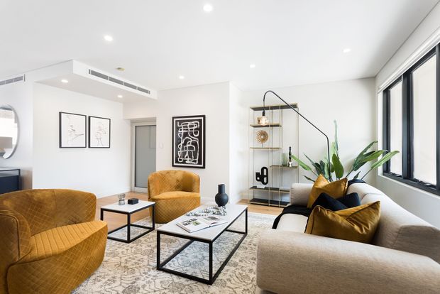 Bright contemporary living room with a large neutral sectional sofa, two mustard upholstered armchairs, nested black metal coffee tables, patterned area rug, sculptural decor, open shelving, framed artwork, indoor plants, and expansive black-framed windows. The room has a clean white palette with warm mustard and wood accents.