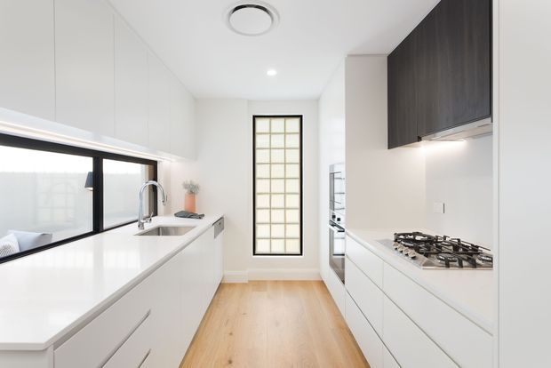 Bright, narrow contemporary galley kitchen with white full-height cabinetry, white stone-look benchtops, a gas cooktop, integrated sink, timber flooring, dark upper cabinetry, black-framed glazing and a tall glass-block window. The space is clean and minimalist with abundant natural light.