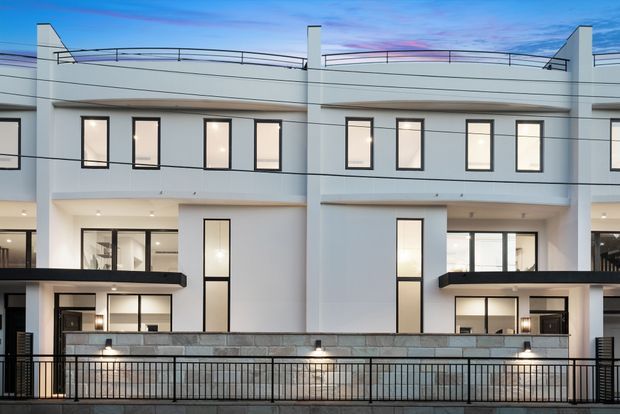 Contemporary multi-storey residential facade with a symmetrical composition, white rendered walls, extensive black-framed glazing, projecting dark balcony awnings, and a stone-clad lower level. Black metal railings run along the foreground, while rooftop cable or wire balustrades and sculptural parapet elements define the upper terrace against a blue evening sky.