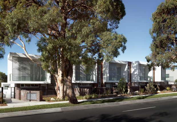 Street-facing view of a contemporary multi-unit residential development with repeated box-like upper-level forms, extensive horizontal external louvres, pale rendered or metal-clad walls, and dark brickwork at the lower levels. Mature eucalyptus trees line the verge and partially screen the building. The site includes a paved footpath, landscaped front setbacks, and a quiet suburban road.