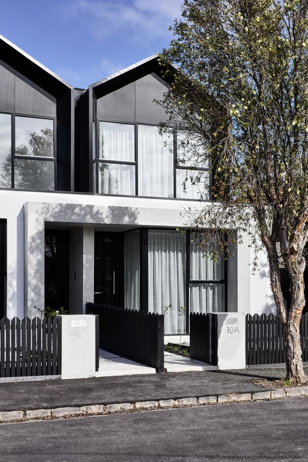 Street-facing view of a contemporary multi-level townhouse with a sharply articulated pitched/gabled roof form, dark charcoal upper facade panels, white masonry or rendered lower facade, large black-framed windows, and a recessed entry. A black picket fence and gate sit in front, with a mature street tree partially obscuring the right side.