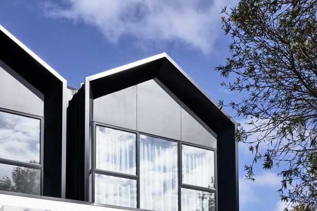 Close-up exterior view of a contemporary multi-level building facade with sharply angled gabled roof forms, dark metal trim, smooth light-grey wall panels, and large window assemblies with sheer curtains. A neighbouring facade is partially visible at left, with a tree and blue sky in the background.
