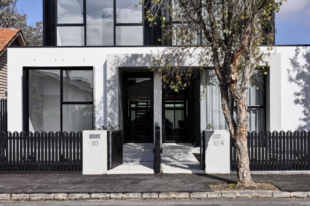 Street-facing exterior of a contemporary multi-level residence with a crisp white rendered facade, tall black-framed glazing, recessed double entry doors, and a dark vertical picket fence. A mature street tree partially screens the entrance, while neighbouring rooflines and the footpath frame the urban residential setting.