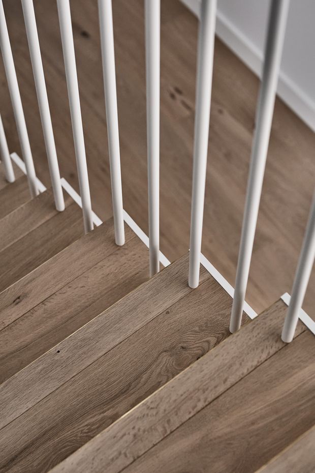 Close-up architectural detail of a staircase with warm natural timber treads and slender white vertical metal balusters. The image focuses on the junction between the stair treads, railing, and adjacent timber flooring, creating a clean, refined contemporary look.