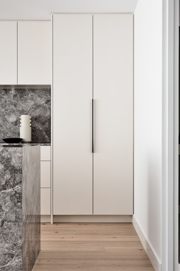 A close-up view of a minimalist contemporary kitchen, focused on tall, handle-accented pale cabinetry and a dark veined stone splashback and island edge. Light timber flooring, white walls, and restrained black and neutral detailing create a clean, refined interior.