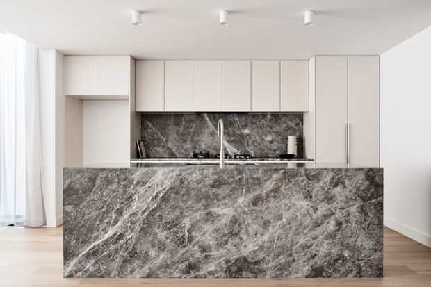 A bright, minimalist contemporary kitchen centered on a large waterfall-edge island clad in dramatic grey veined stone. The kitchen has streamlined off-white flat-panel cabinetry, a matching stone splashback, integrated appliances, a gas cooktop, and a tall brushed-metal tap. Light timber flooring and sheer curtains add warmth to the otherwise monochromatic space, with simple ceiling-mounted spotlights providing illumination.