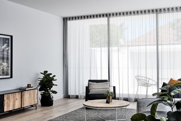 Bright, contemporary living room with white walls, pale timber flooring, full-height sheer curtains, and expansive glazing. The room contains a low timber media cabinet, black-framed lounge chair, grey sofa, round timber coffee table, patterned rug, indoor plants, and framed wall art. The neutral palette and abundant filtered daylight create an airy, calm atmosphere.