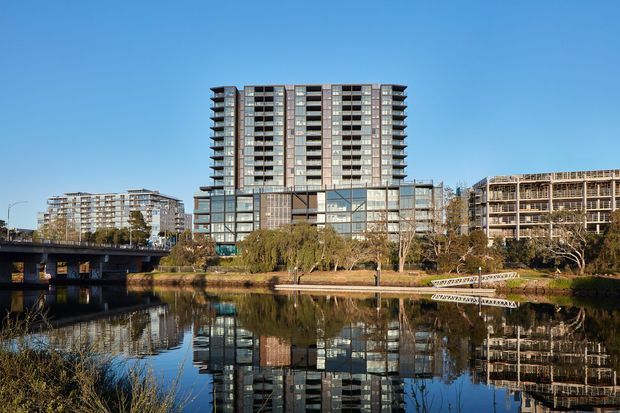 Exterior architectural view of a contemporary multi-storey apartment or mixed-use development beside a calm reflective waterway. The central tower has a regular grid facade with extensive glazing, balconies, and dark-and-light vertical elements, flanked by lower and taller surrounding buildings. A pedestrian path, landscaped riverbank, bridge, and strong building reflections are visible under clear daylight.