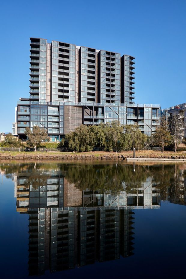 Exterior view of a contemporary multi-storey apartment building beside a calm body of water, with extensive glazed facades, projecting balconies, dark metal framing and varied tower volumes. The building and surrounding trees are reflected prominently in the water under a clear blue sky.
