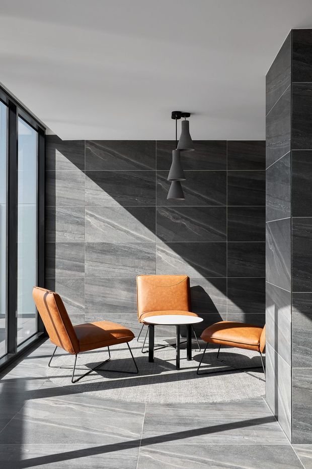 A compact, sunlit modern lounge or sitting area with floor-to-ceiling black-framed glazing on the left, large dark grey stone-look tiled walls and flooring, and a matching tiled structural pier on the right. Three cognac-brown leather lounge chairs surround a small round white table, with three matte-black pendant lights hanging above. Strong diagonal sunlight and shadows create a dramatic, refined atmosphere.
