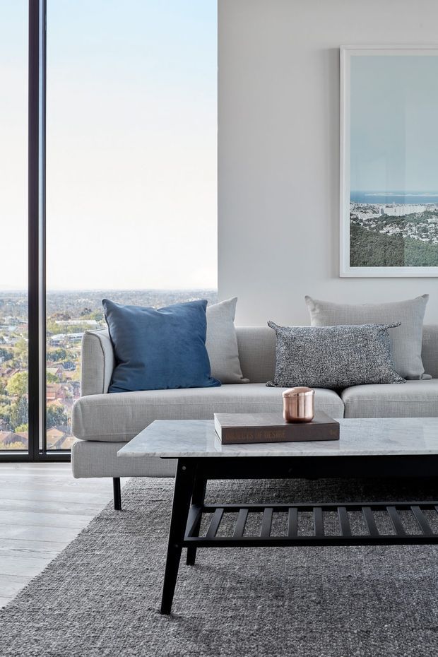 A bright, contemporary living room with a large floor-to-ceiling window overlooking a leafy urban landscape and distant coastline. The room features a light grey sectional sofa with blue and neutral cushions, a pale marble coffee table with a black slatted lower frame, and a textured grey rug. A framed coastal landscape photograph hangs on the light wall.