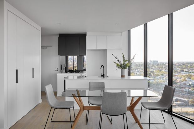 A bright, contemporary open-plan kitchen and dining area in a high-rise apartment, with white floor-to-ceiling cabinetry, a white island with integrated sink, dark feature cabinetry, pale timber flooring, and a glass dining table with upholstered chairs. Full-height black-framed glazing provides expansive city views and abundant natural light.