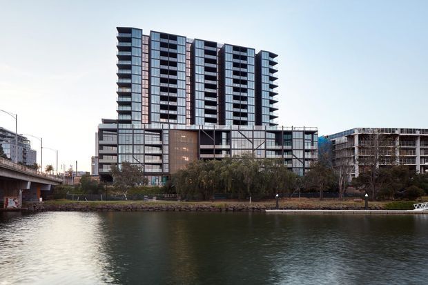 Exterior view across a body of water toward a contemporary multi-storey apartment or mixed-use building. The main structure has a tall, rectilinear tower with extensive dark glazing, projecting balconies, vertical window bands, and a lower podium composed of glass and dark metal panels. Adjacent urban buildings and an elevated roadway are visible, with landscaped trees along the waterfront.