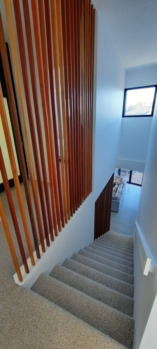 A tall interior stairway viewed from the upper landing, with grey carpeted stairs descending toward a lower living area. A prominent vertical timber slat screen lines the stair opening, contrasting with white walls and a black-framed window that brings in daylight. The lower level includes a partial view of a light-coloured sofa and tiled or polished flooring.
