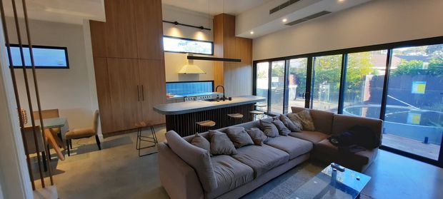 Open-plan contemporary living area with a large sectional sofa in the foreground, a compact kitchen and island behind it, timber-lined cabinetry and feature wall, black-framed full-height glazing, and polished concrete flooring. The space has a refined, warm-modern fit-out with integrated lighting and a strong connection to an outdoor terrace.