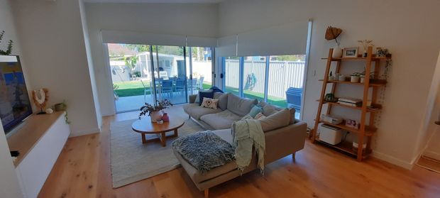 Bright open-plan living room with warm timber flooring, a large light-grey sectional sofa, round wooden coffee table, neutral rug, and open timber shelving. Full-height glazing and sliding doors open to a fenced backyard and patio, creating an airy indoor-outdoor connection.