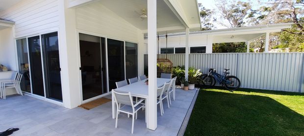 Bright covered outdoor entertaining area adjoining a contemporary white-clad home, with a paved patio, dining setting, outdoor lounge furniture, a small lawn, and a lightweight carport-like structure with bicycles at the rear. The architecture features white horizontal cladding, slender posts, and a shallow skillion-style roof form.