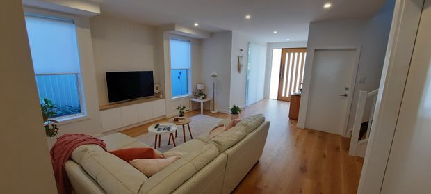 Bright contemporary living room viewed from the rear, with a large cream sectional sofa, wall-mounted television on built-in timber joinery, coffee tables, light neutral walls, recessed downlights, and warm timber flooring. The entry hall and stair are visible in the background, with daylight entering through windows and a glazed front door.