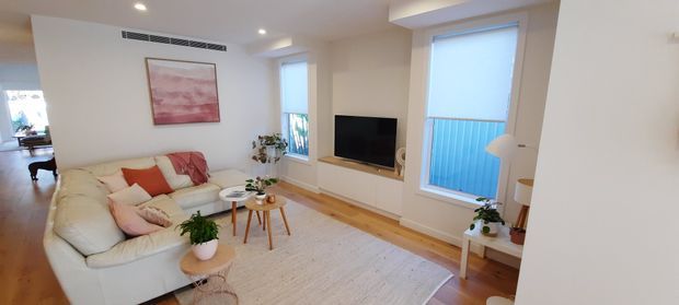 Bright, compact contemporary living room with a cream sectional sofa, pale area rug, nested round coffee tables, timber flooring, recessed lighting, and a wall-mounted television set within a light-toned built-in surround. Large windows bring in daylight, with indoor plants and restrained neutral furnishings creating a warm, inviting atmosphere.