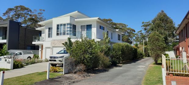 Street-level exterior view of a contemporary multi-level white-clad residence with a low-pitched roof, projecting balcony, large windows, attached garage, driveway, and landscaped front garden. Neighbouring homes and mature trees are visible in the suburban setting.