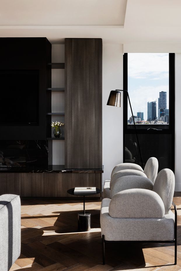 A contemporary living room in a high-rise urban apartment, featuring a dark timber entertainment wall with integrated television and shelving, a tall timber storage cabinet, black stone-look joinery, sculptural light grey upholstered lounge chairs, and a small black side table. Warm herringbone timber flooring contrasts with white walls and black-framed glazing, which overlooks a dense city skyline.