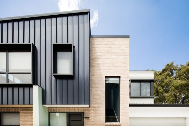 Contemporary multi-level residential facade with dark vertically ribbed metal cladding, pale brick masonry, projecting window surrounds, large glazed openings, and a partially visible integrated garage. The clean geometric composition is set against a blue sky and surrounding trees.