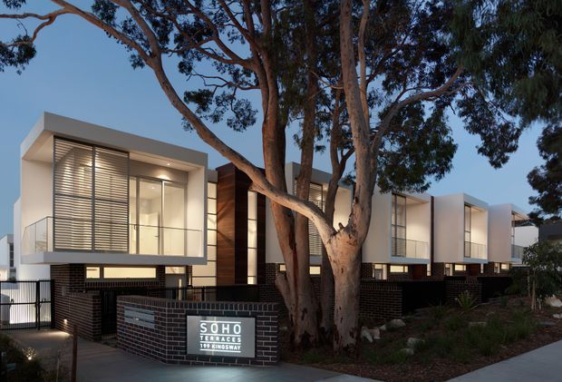 Twilight exterior view of a contemporary multi-unit residential development, with projecting box-like apartments, pale rendered facades, extensive glazing, horizontal external blinds, dark brickwork, timber-clad vertical accents, and landscaped native trees. A sign identifies the development as Soho Terrace. Large established eucalyptus trees dominate the foreground.