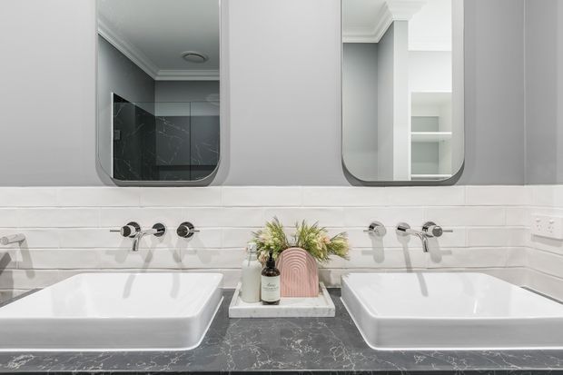 A contemporary double-vanity bathroom with two white vessel basins set on a dark marble-look benchtop. Twin wall-mounted chrome tap sets sit beneath two rounded rectangular mirrors, with white horizontal wall tiles, grey painted walls, and built-in shelving visible in the reflections.