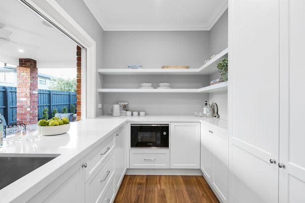 Bright white U-shaped kitchen with shaker-style cabinetry, a white benchtop, integrated microwave, open display shelving, stainless-steel sink, and warm timber flooring. Large open glazing connects the kitchen to a covered outdoor area and fenced yard. The room has a clean, traditional-leaning design with abundant natural light.