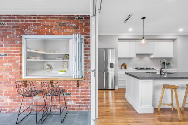 Split view of a renovated brick home showing an exposed red-brick entertaining area with a servery window, metal bar stools, and grey tiled flooring beside a bright white kitchen with shaker-style cabinetry, stainless-steel refrigerator, stone island benchtop, timber flooring, pendant lighting, and timber stools.