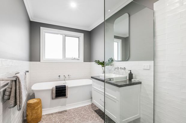 Bright contemporary bathroom with a freestanding white bathtub beneath a window, a white vanity with dark stone benchtop, large mirror, glass shower enclosure, white subway wall tiles, grey painted upper walls, decorative patterned floor tiles, and simple timber and textile accents.