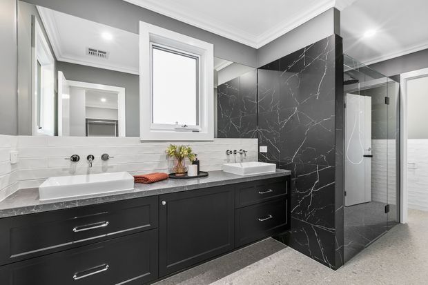 Contemporary bathroom with a long double vanity in dark cabinetry, white basins, a dark marble-look tiled feature wall, large mirror, and a frameless glass shower enclosure. The room has light grey walls, white subway-style wall tiling, polished chrome fixtures, and a light speckled floor.