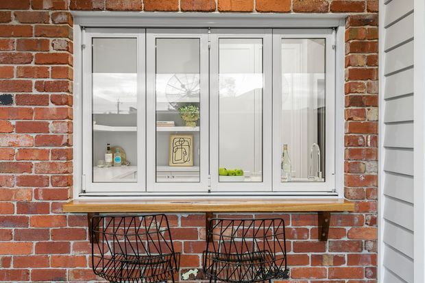Exterior view of a brick facade with a large white-framed multi-panel window. The window reflects the surrounding landscape and reveals a bright interior with shelving, small objects, and a sink tap. Two black wire-frame stools sit beneath the projecting sill, with white horizontal cladding visible at the right edge.