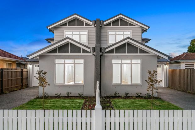 Front exterior view of a symmetrical contemporary duplex or pair of attached homes, featuring grey weatherboard cladding, white trim, multiple gabled roof forms, prominent central downpipes, large white-framed windows, separate paved driveways, small front lawns, timber side fencing, and a white picket fence.