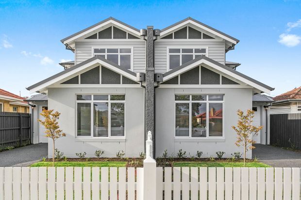 Front exterior view of a symmetrical two-storey duplex or townhouse-style residence, featuring grey horizontal weatherboard cladding, multiple pitched gables, white trim, large white-framed windows, a central textured vertical chimney or dividing feature, front paths, small planted gardens, and a white picket fence.