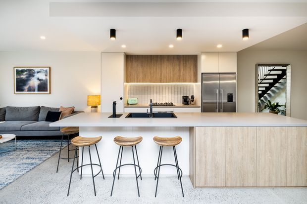 A bright, open-plan contemporary kitchen and living area. The kitchen features a large light-toned island with integrated sink and seating for three, pale timber cabinetry, a wood-look feature wall, stainless-steel appliances, and a tiled backsplash. A grey sectional sofa and rug occupy the adjoining living area, while a staircase is visible through an opening at the rear.